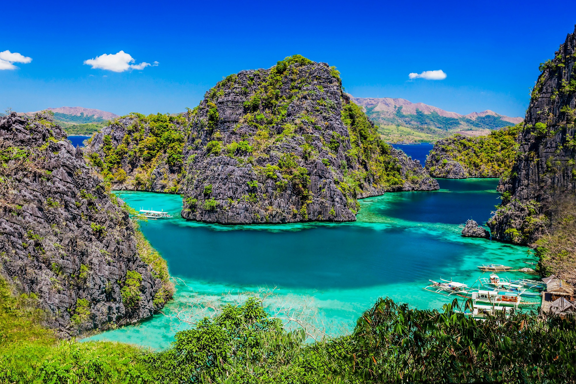 Hohe, teil bewachsene Felsen ragen aus dem türkisem Wasser in den blauen Himmel. Ein paar kleine Boote liegen vor Anker.