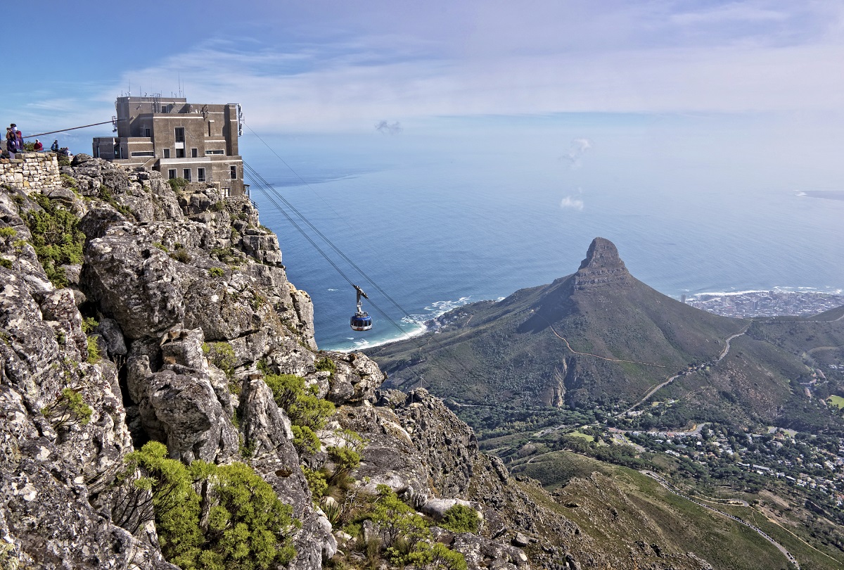 Eine Seilbahn fährt auf den felsigen Tafelberg zu. Unten liegt eine grüne Landschaft mit dem markanten Lion’s Head, dahinter das weite blaue Meer unter einem leicht bewölkten Himmel.