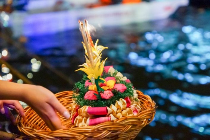 Hand hält einen geflochtenen Korb mit buntem Blumenarrangement und brennenden Kerzen darüber. Der Hintergrund zeigt verschwommene Wasseroberfläche mit Lichtern und Reflexionen bei Nacht.