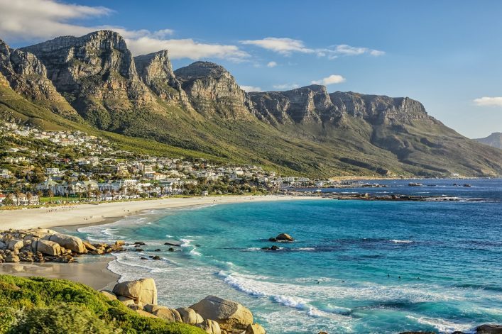 Weißer Sandstrand mit türkisfarbenem Meer im Vordergrund, dahinter eine Stadtlandschaft und imposante Berge unter strahlend blauem Himmel.
