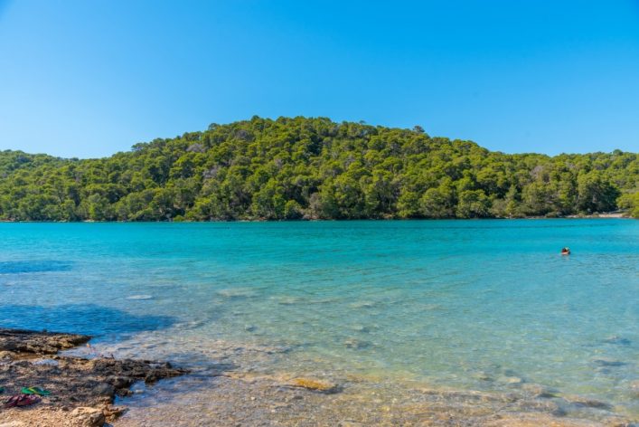 Klares, türkisblaues Wasser eines Sees im Vordergrund, von felsigem Ufer gesäumt. Im Hintergrund erhebt sich ein bewaldeter Hügel unter klarem blauem Himmel. Einzelner Schwimmer im Wasser rechts.