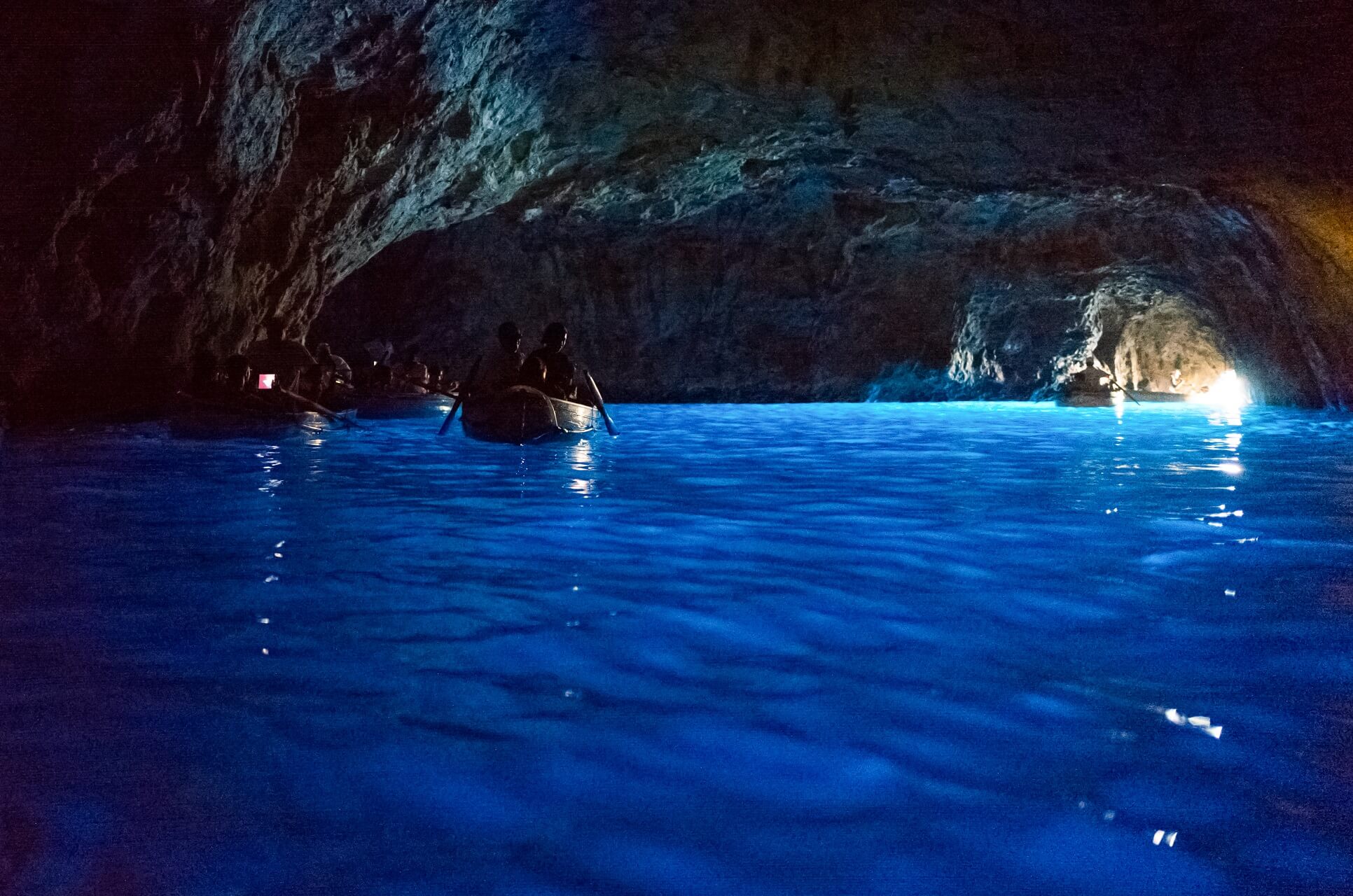 Boote mit Menschen in einer dunklen, natürlichen Höhle; das Wasser leuchtet intensiv blau durch Lichteinfall von rechts hinten. Die Höhlenwände haben unregelmäßige Strukturen in Grautönen.