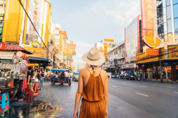 Frau in orange Kleidung mit Hut betrachtet die lebendige Khaosan Road in Bangkok; Straße mit Schildern und Tuk-Tuk. Packliste für Thailand im Gepäck.