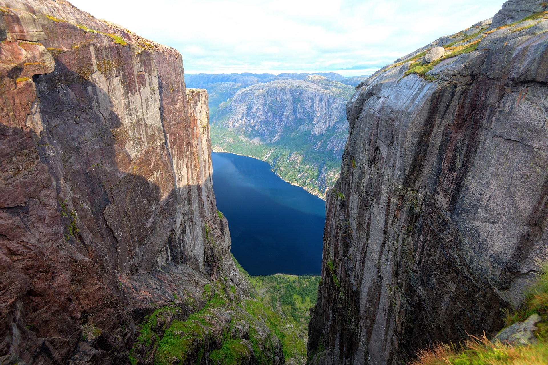 Tiefe Felsschlucht mit steilen, dunklen Felswänden, am Grund ein dunkelblauer Fjord, umgeben von einer weiten Berglandschaft.