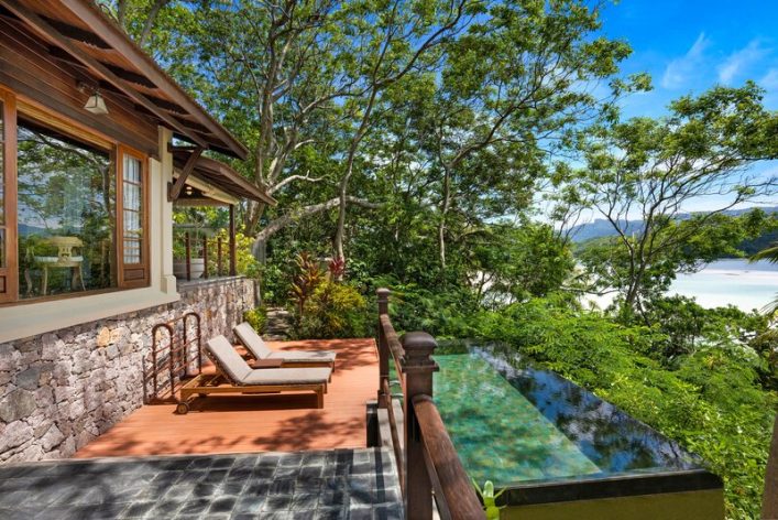 Holzterrasse mit zwei Liegestühlen neben einem Infinity-Pool, umgeben von üppiger Vegetation. Links ein Steinhaus mit großen Fenstern. Im Hintergrund blauer Himmel und Meerblicke durch die Bäume.