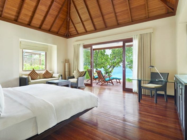 Geräumiges Hotelzimmer mit Holzboden, einem großen Bett links, einer Sitzgruppe und einem Schreibtisch rechts. Große Fenster bieten Blick auf Meer und grüne Vegetation. Schiebetüren führen auf die Terrasse.