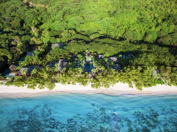 Tropische Hotelanlage eingebettet in dichten, grünen Dschungel, der sich bis zum weißen Sandstrand erstreckt. Dahinter leuchtet das türkisfarbene Meer. Gebäude sind umgeben von Palmen und Vegetation.