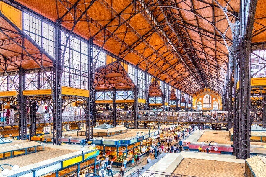 Luftaufnahme einer geräumigen Markthalle in Budapest mit hohen Stahlträgern und einem Holzdach. Unten reihen sich bunte Marktstände aneinander, während sich Besucher durch die Gänge bewegen.