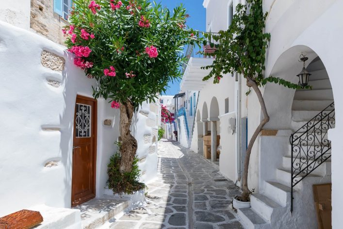 Enge, weiß getünchte Gasse auf der Insel Paros, gesäumt von leuchtend pinken Blumen und blühenden Bäumen. Links eine Holztür, rechts eine gewundene Treppe und geschmiedete Geländer, blauer Himmel.