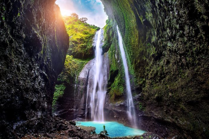 Madakaripura-Wasserfall in Java stürzt von steilen, bewachsenen Felsen in ein türkisfarbenes Becken. Sonnenlicht scheint von oben, während eine Person unten steht und die Szene betrachtet.