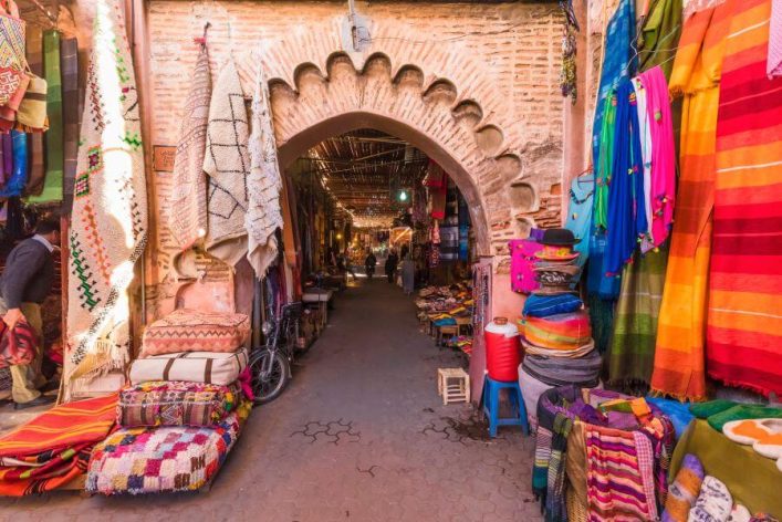 Bunter Markt in Marrakesch unter einem steinernen Bogen mit Stoffen und Kissen. Besuche früh für weniger Trubel und bessere Deals.