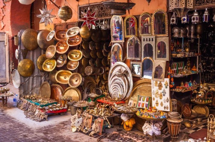 Farbenprächtiger Souk in Marrakesch: Metallteller und Lampen stapeln sich vor einer Wand, bunte Teppiche und Spiegel ergänzen die Szenerie, ein Hauch von Handwerkskunst und Kultur.