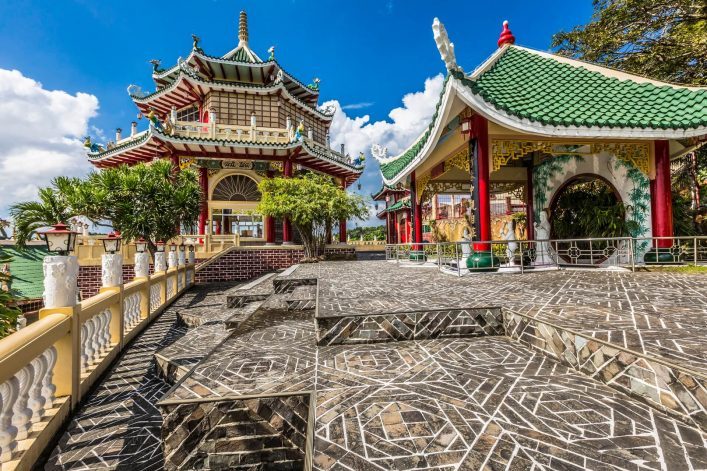 Taoistischer Tempel in Cebu mit grünen Dächern, verzierten Pagoden und Drachen-Skulpturen. Links große Pagode, rechts kleiner Pavillon, dazwischen Steinplattenweg. Bäume und blauer Himmel im Hintergrund.