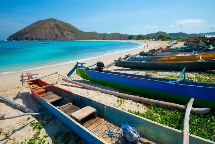Mehrere bunte Fischerboote liegen auf einem sandigen Strand. Dahinter erstreckt sich das türkisfarbene Meer mit einer bewaldeten Hügelkette im Hintergrund unter klarem Himmel.