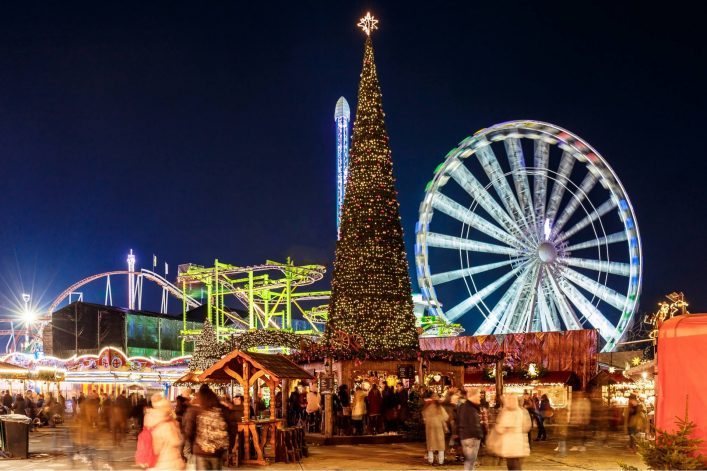 Weihnachtsmarkt mit großem, beleuchtetem Weihnachtsbaum zentral, Riesenrad rechts im Hintergrund, bunte Fahrgeschäfte links, Menschenmengen im Vordergrund. Festliche Lichter erhellen die Szene.