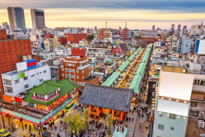 Blick auf den belebten Asakusa-Bezirk in Tokio. In der Mitte verläuft eine lange, überdachte Einkaufsstraße zu einem Tempel. Umgeben von bunten Gebäuden, im Hintergrund moderne Hochhäuser bei Sonnenuntergang.