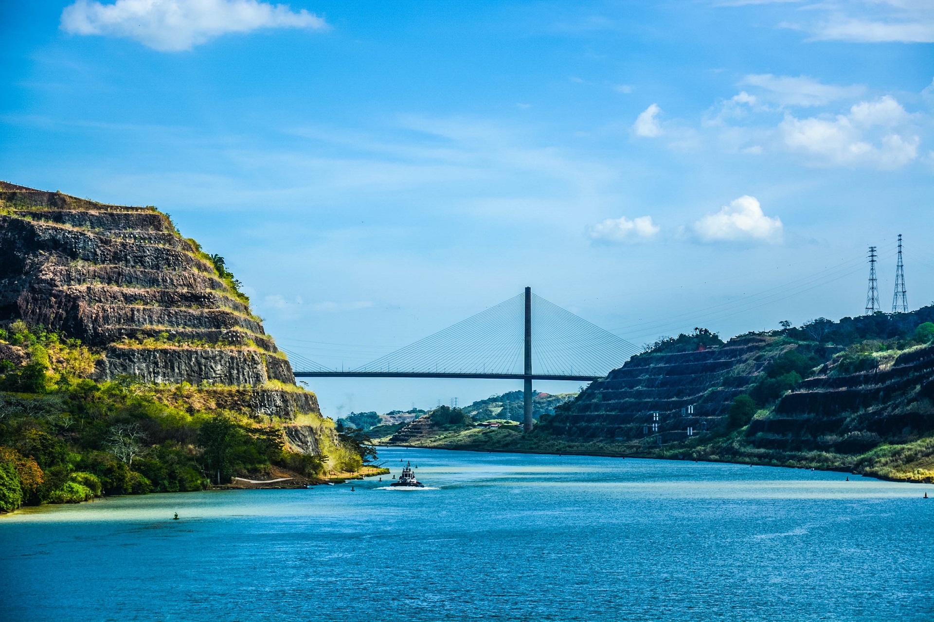 Blick auf den Panama-Kanal mit einer Brücke im Hintergrund, flankiert von terrassierten Hügeln und blauem Himmel.
