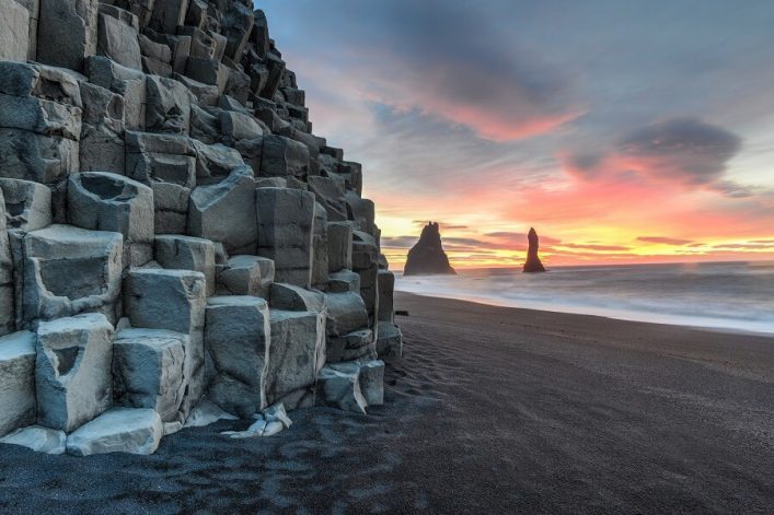 Basaltsäulen am linken Rand, schwarzer Sandstrand in der Mitte, Meer und zwei Felsnadeln im rechten Drittel, bunter Sonnenuntergangshimmel.