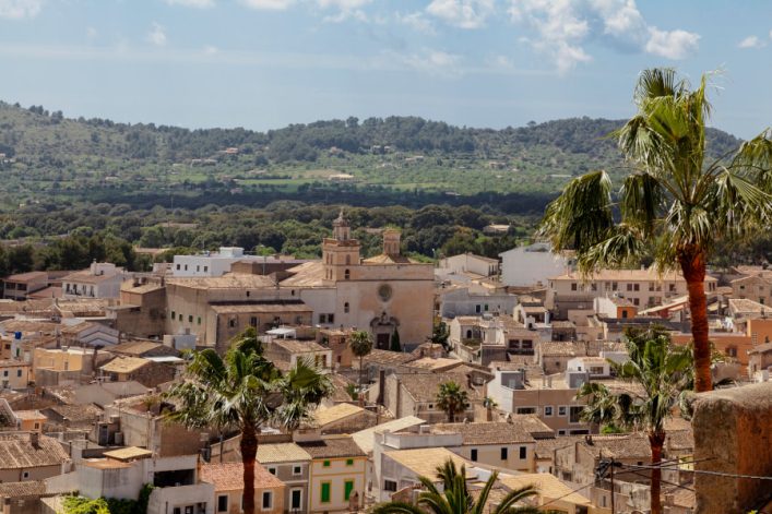 Blick über die Dächer der Stadt Artà auf Mallorca mit einer Kirche in der Bildmitte. Im Vordergrund befinden sich hohe Palmen. Der Hintergrund zeigt bewaldete Hügel unter einem blauen Himmel.