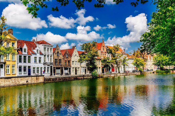 Buntes Reihenhauspanorama an einem ruhigen Kanal in Brügge, gesäumt von Bäumen unter blau-weißem Himmel.