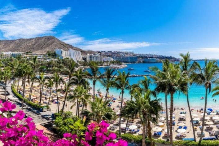 Türkisblaues Meer mit Sandstrand, gesäumt von Palmen. Menschen entspannen unter Sonnenschirmen. Im Hintergrund Berge und weiße Gebäude, darüber blauer Himmel. Im Vordergrund rosa Blumen.