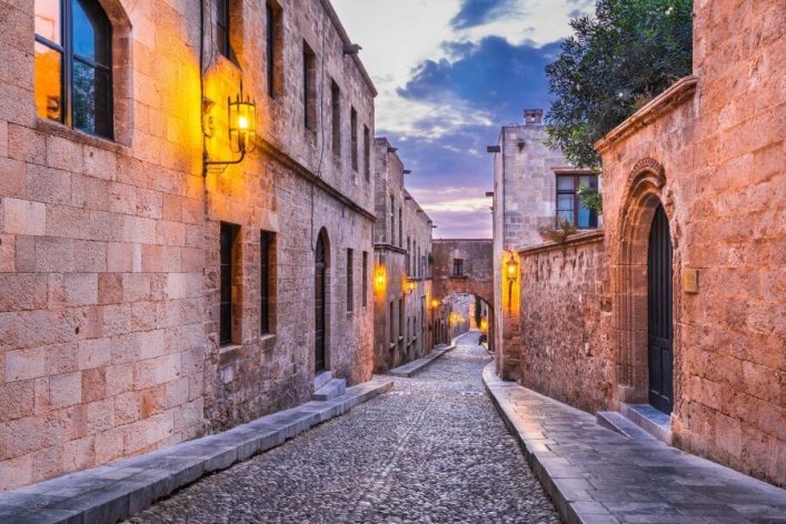 Historische, gepflasterte Gasse in Rhodos-Stadt bei Dämmerung, flankiert von steinernen Bauten mit beleuchteten Laternen und einer Bogenbrücke im Hintergrund.