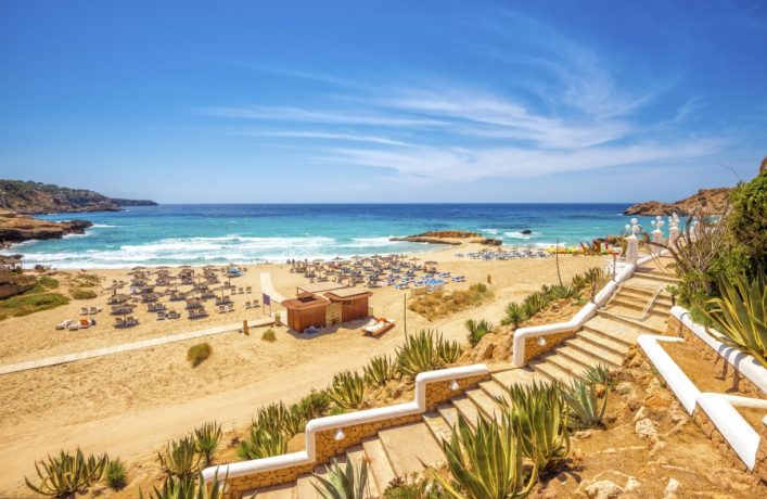 Treppen führen zur Cala Tarida auf Ibiza. Unten sind Sonnenschirme und Liegestühle in Reih und Glied auf dem goldenen Sand angeordnet, mit türkisfarbenem Meer und blauem Himmel im Hintergrund.