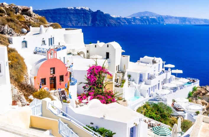 Weiß getünchte Häuser mit blauen Akzenten auf Klippen von Santorini, darüber blauer Himmel, unten das ruhige Meer. Farbtupfer: rosafarbene Blüten, roter Windmühlendach.