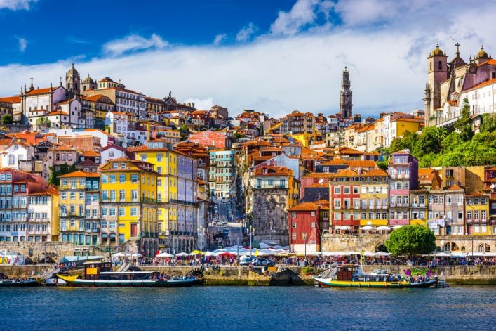 Bunte Häuser mit roten Dächern steigen einen Hügel hinauf, dominieren die Altstadt von Porto. Im Vordergrund fließt ein Fluss, an dem Boote liegen. Der Himmel ist teils bewölkt.