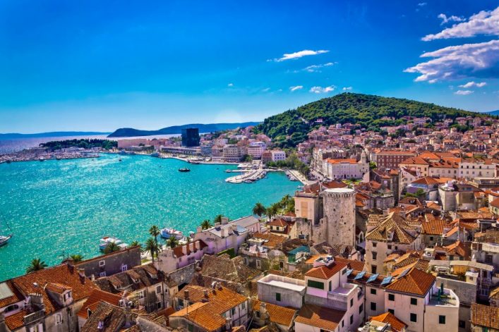 Blick von oben auf die Küste von Split, Kroatien. Im Vordergrund historische Gebäude mit roten Dächern, dahinter das türkisfarbene Meer. Im Hintergrund erhebt sich der grüne Marjan-Hügel.