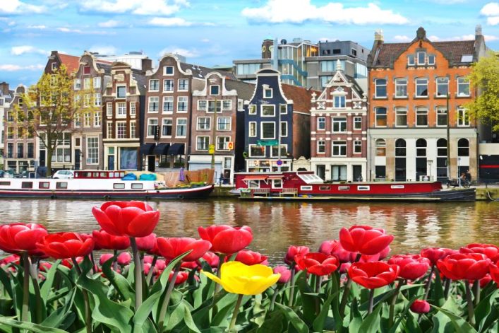 Im Vordergrund leuchtende rote Tulpen, eine gelbe sticht hervor. Dahinter fließt ein Kanal, gesäumt von bunten, schmalen Häusern und Hausbooten unter einem blauen, bewölkten Himmel.