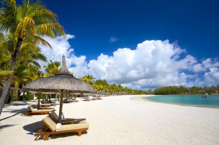 Strand mit weißen Liegen unter Strohdächern, Palmen links, feiner Sand, türkisblaues Meer rechts. Am Horizont ein klarer, blauer Himmel mit einigen weißen Wolken.