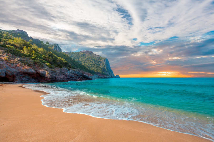 Goldener Sandstrand führt zu türkisfarbenem Meer, flankiert von grünen Klippen unter einem malerischen Sonnenuntergang, ideal für Strandurlaub im April.