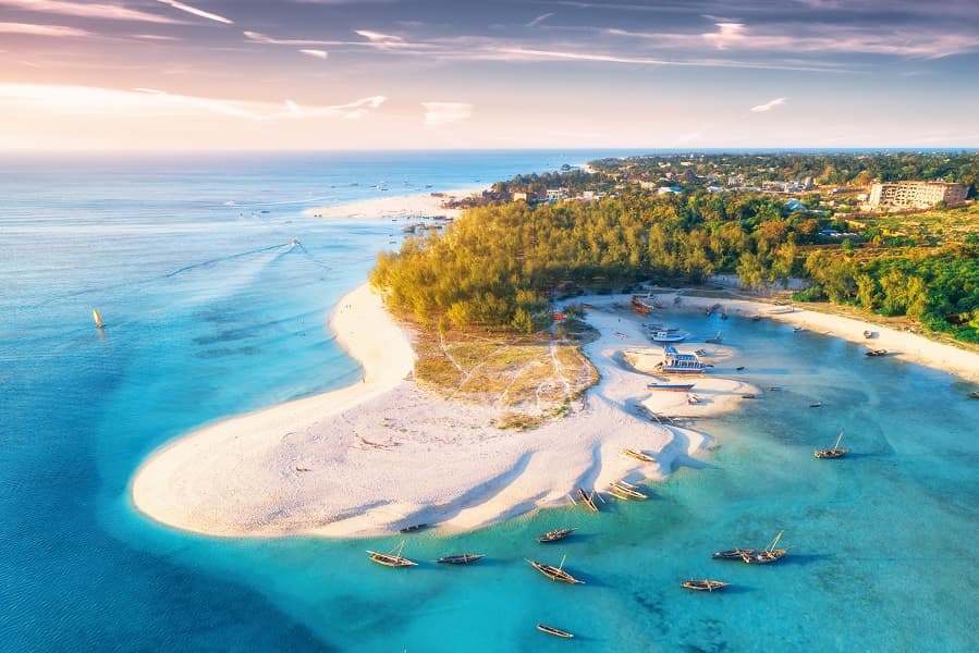 Eine Drohnenaufnahme des runden, perlweißen Strandes auf Sansibar. Im Wasser liegen braune, kleine Holzboote im türkisfarbenen, glasklaren Wasser. Der Strand geht über in dichten, sattgrünen Palmenwald.