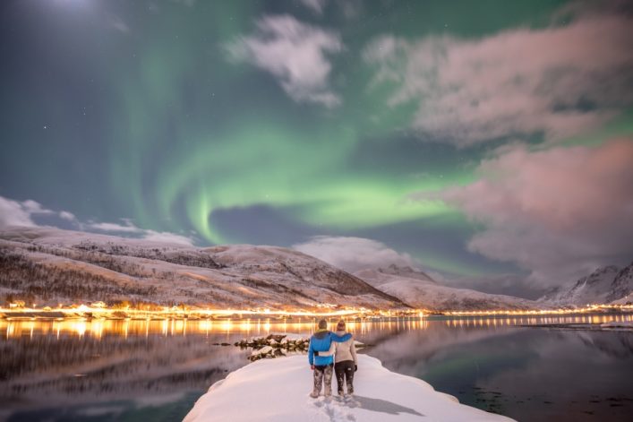 Ein Paar steht auf verschneitem Ufer, umarmt, blickt auf Nordlichter über einer norwegischen Spiegelwasser-Bucht mit beleuchteten Häusern.