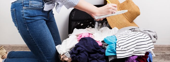 Eine kniende Person in Jeans packt einen überfüllten Koffer mit bunter Kleidung, einem Strohhut und einem Streifenheft. Der Koffer liegt geöffnet auf einem Holzboden.