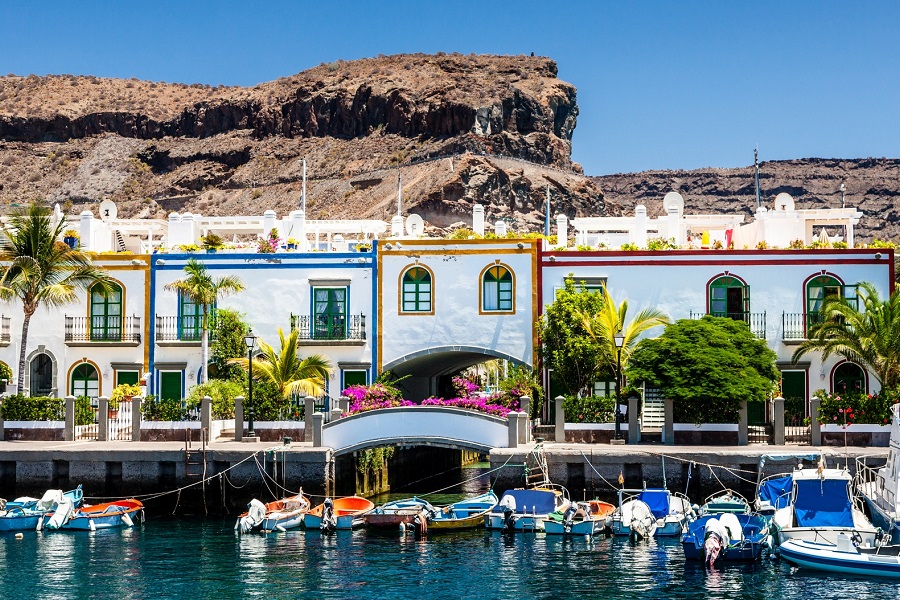Bunte, zweistöckige Häuser am Hafen von Puerto de Mogán, Gran Canaria. Palmen und blühende Pflanzen davor, Boote im Wasser spiegeln sich. Perfekter Ort für einen entspannten Familienurlaub.