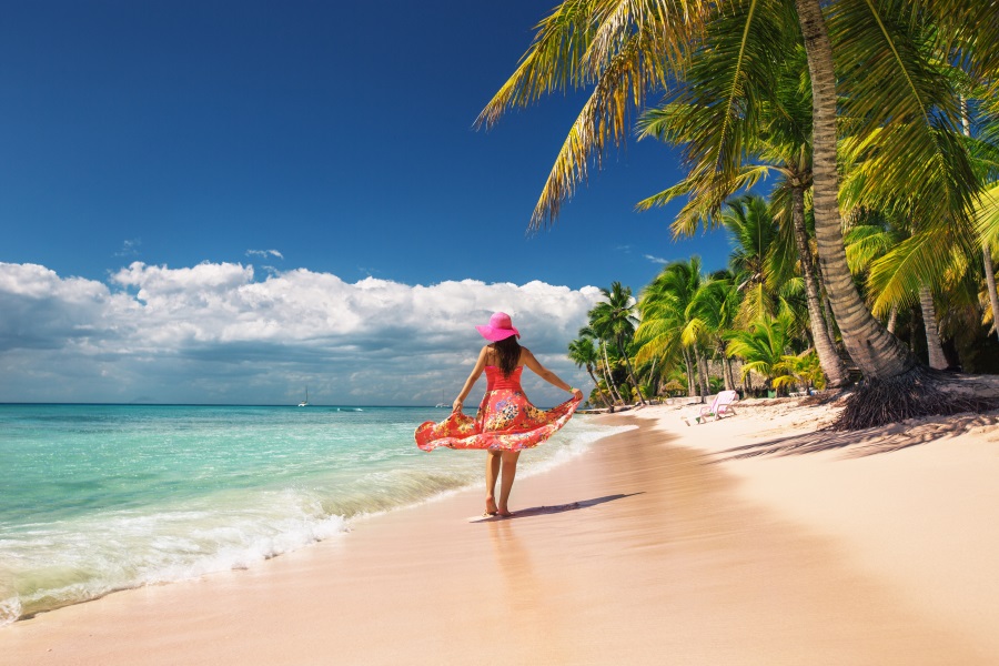 Frau mit rotem Kleid und rosa Hut läuft an einem karibischen Strand entlang, ideal für Fernreisen im Mai, umgeben von Palmen und türkisem Meer.