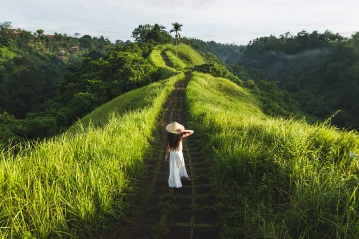 Frau in weißem Kleid und Strohhut spaziert auf steinigem Pfad inmitten grüner Reisterrassen in Ubud, Bali, mit sanften Hügeln im Hintergrund.