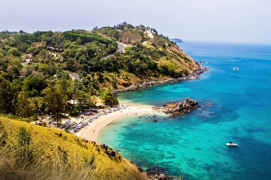 Feinsandiger Strand mit Sonnenschirmen und Liegen, eingerahmt von grüner Vegetation und Hügeln. Das kristallklare, türkisfarbene Meer zieht sich bis zum Horizont, wo Boote auf dem Wasser treiben.