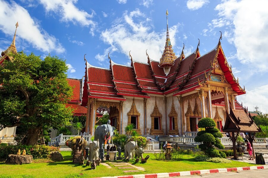 Buddhistischer Tempel in Thailand mit roten Dächern und goldenen Verzierungen, umgeben von Bäumen und Elefantenstatuen, unter strahlend blauem Himmel mit einigen Wolken.