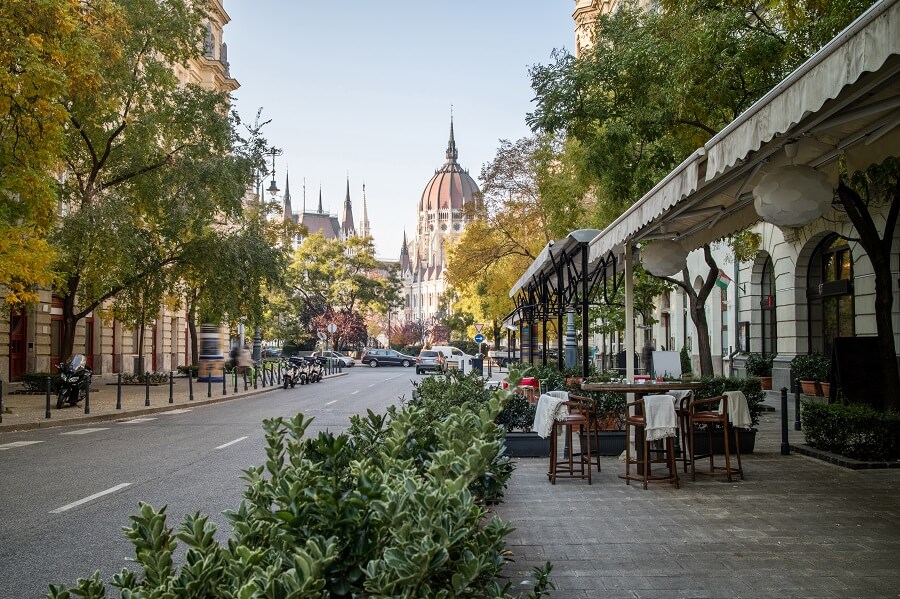 Durch die Mitte des Bildes läuft eine Straße. Am Ende der Straße ist das Parlament von Budapest zu erkennen. Links und rechts von der Straße stehen grüne Bäume. Am rechten Bildrand sind Cafés zu erkennen.