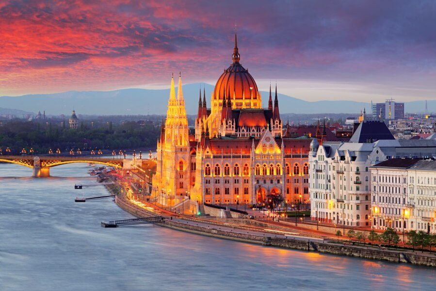 Das Parlament von Budapest aus der Luft fotografiert. Es dämmert bereits, weshalb der Himmel lila-rot leuchtet. Das Parlament schein in einem hellen gelb-roten Licht und sticht durch die vielen Türme und die große runde Kuppel in der Mitte direkt ins Auge. Vor dem Parlament läuft ein Fluss entlang.