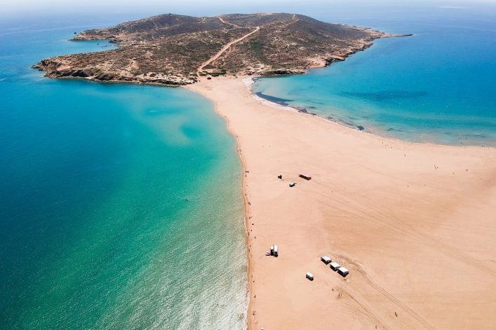 Eine schmale Landzunge trennt zwei blaue Meeresabschnitte, verbindet eine karge Insel mit dem Festland, einige Fahrzeuge am Strand.