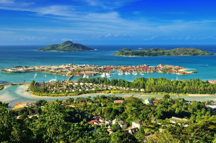 Panoramablick von grünen Hügeln auf eine tropische Bucht mit einem Dorf auf den Seychellen, umgeben von türkisblauem Meer und Inseln.
