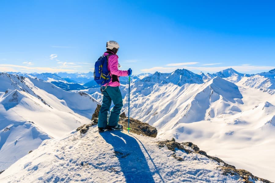Auf diesem Bild steht eine Frau auf einem Berg in den Österreicher Alpen und blickt auf eine schneebedeckte Alpenlandschaft. Sie trägt einen Skihelm, eine pinke Skijacke, Skischuhe und trägt Skistöcke bei sich. Der Himmel ist blau und die Sicht klar.