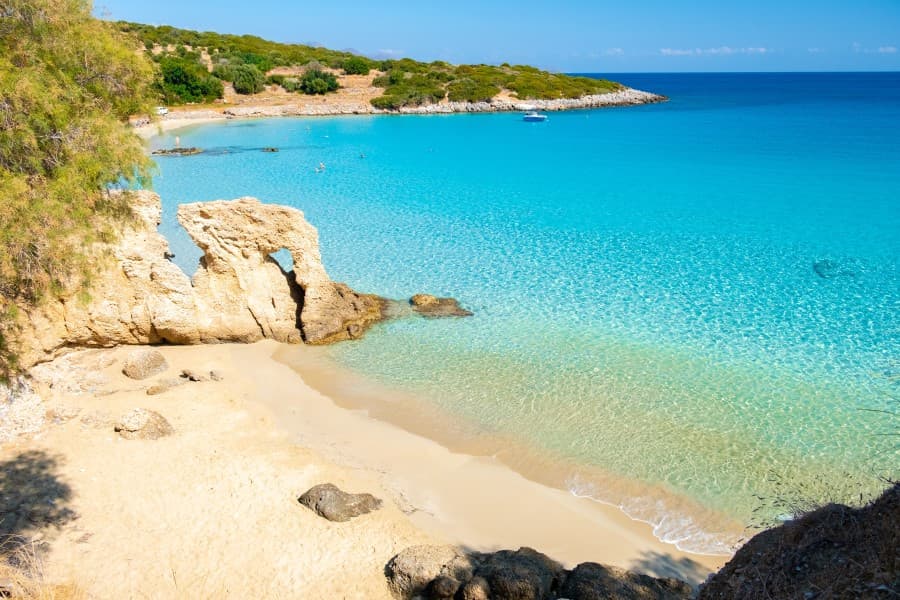 Türkisfarbenes Meer umspült eine kleine Sandbucht auf Kreta, umrahmt von felsigen Klippen und spärlicher Vegetation, unter klarem blauen Himmel.