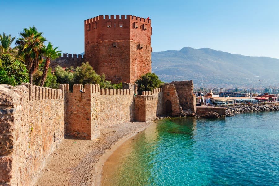 Der Rote Turm in Alanya, umgeben von Steinmauern, steht neben dem türkisblauen Meer; im Hintergrund erheben sich Bergketten.