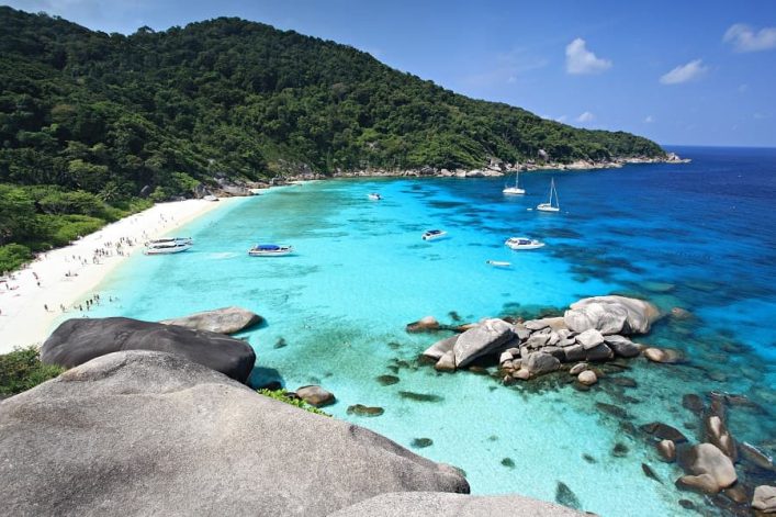 Tropische Bucht in Thailand mit türkisblauem Wasser, weißem Sandstrand, Booten und üppiger grüner Vegetation an den Hängen.