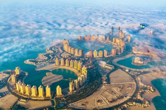 Luftaufnahme von Doha, Katar, einer der sonnigsten Orte der Welt, umgeben von blauem Wasser, Wolken und Hochhäusern, die im Sonnenlicht leuchten.